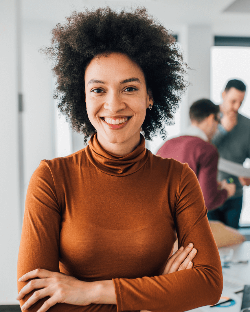 Eine Frau steht mit verschränkten Armen in einem Büro und lächelt, während im Hintergrund Kollegen arbeiten.